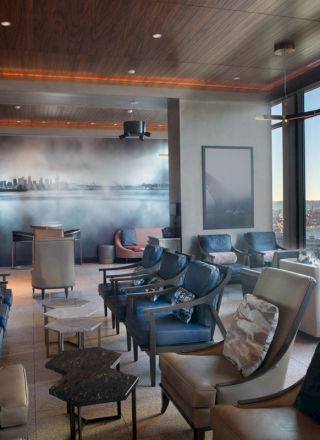 A modern lounge area with multiple chairs, tables, large windows with an ocean view, and a wall mural depicting a cityscape in the background.