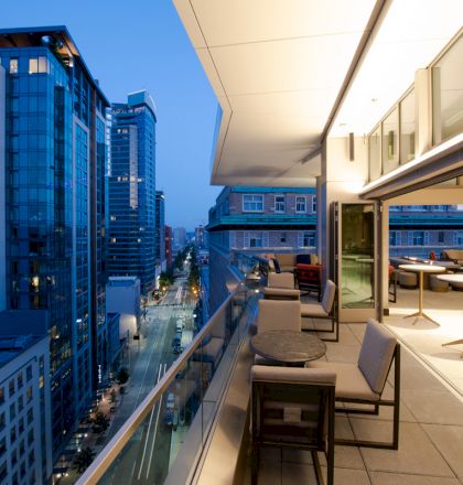A modern rooftop patio with tables and chairs overlooks a bustling city street, surrounded by tall, illuminated buildings at twilight.