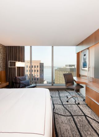 A modern hotel room with a large bed, wall-mounted TV, desk, and chair, featuring cityscape views through the floor-to-ceiling windows.