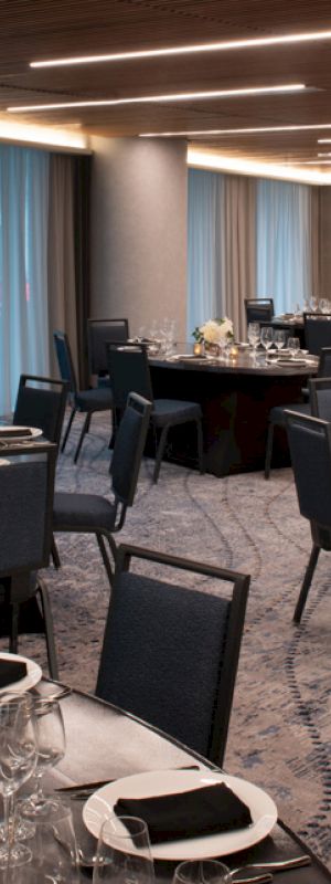 The image shows an elegantly set dining room with round tables, chairs, and dinnerware for an event. The room has dim lighting and modern design.