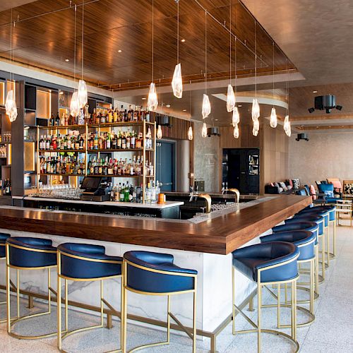This image shows a modern bar with high chairs, a stocked liquor shelf, hanging lights, and lounge seating in the background.