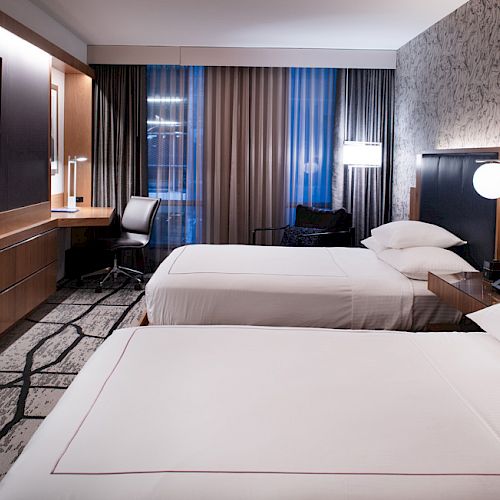 This image shows a modern hotel room with two beds, a TV on the wall, a desk with a chair, and stylish decor including patterned wallpaper and lighting.