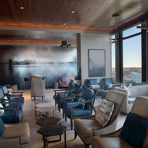 This image shows a modern lounge with comfortable seating, large windows offering a scenic view of the city and a Ferris wheel by the water.