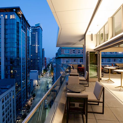 An upscale, modern balcony with seating, tables, and views of a cityscape at dusk. High-rise buildings are visible along the street.
