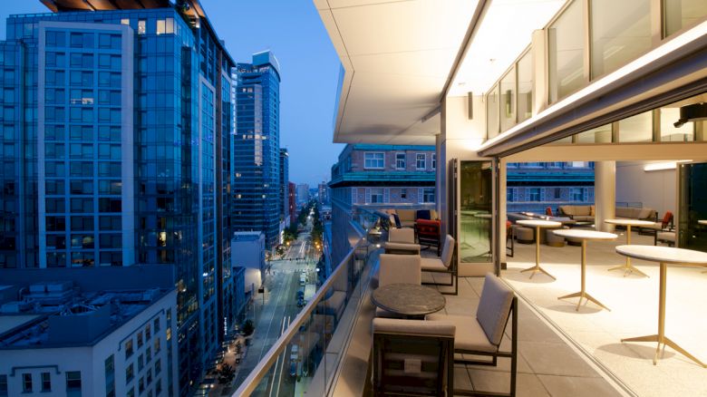 A modern balcony overlooking a city street at dusk, with tables and chairs for seating and tall buildings lining both sides of the street.