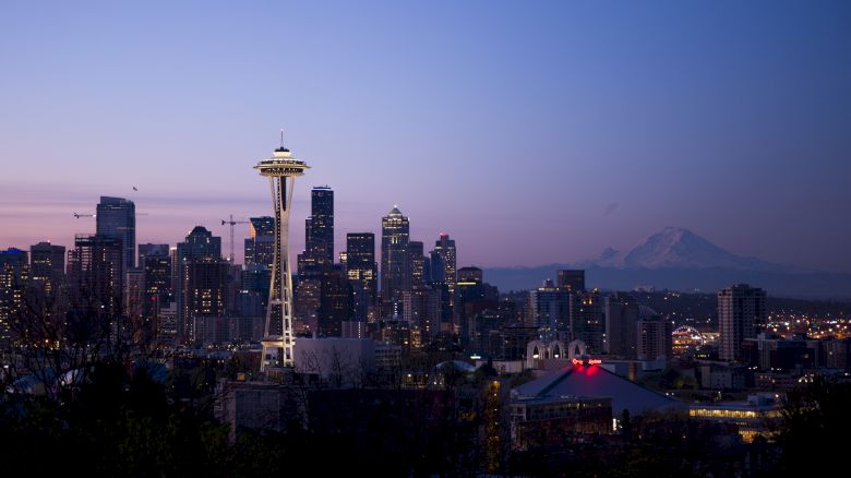 The image shows the Seattle skyline at dusk, featuring the Space Needle with Mount Rainier visible in the background and city lights starting to illuminate.