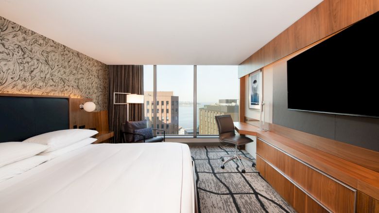 A modern hotel room with a large bed, desk, chair, flat-screen TV, and floor-to-ceiling windows offering a city view with buildings.