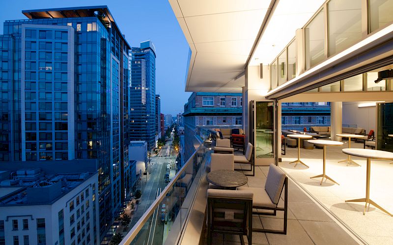 The image shows a modern cityscape at twilight with high-rise buildings and an open balcony featuring seating and tables along the railing.