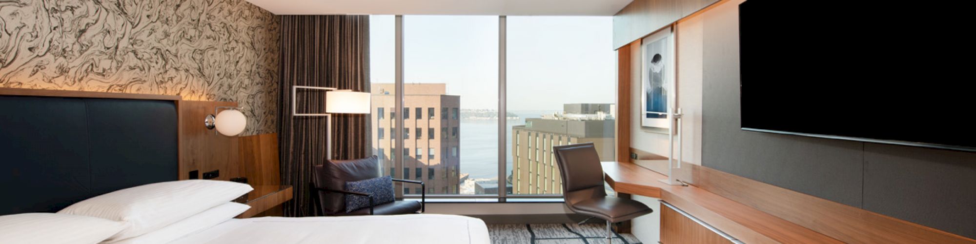 A modern hotel room with a large bed, desk, chair, flat-screen TV, and large windows offering a city view, plus contemporary decor.