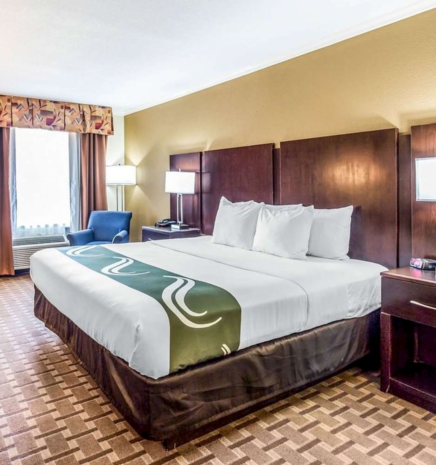 The image shows a neatly arranged hotel room with a double bed, two nightstands, two lamps, a desk, and a chair near the window, well-lit and clean.