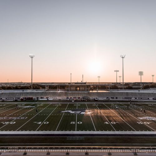 This image shows an empty football stadium at sunset with lights on and a clear sky, casting long shadows across the field.