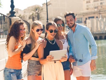 A group of five people, three women and two men, are gathered outdoors by the water, looking at a tablet and smiling.