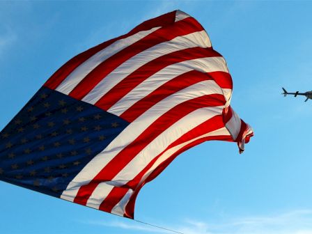 A large American flag is being flown in the sky, with a helicopter visible in the background against a blue sky.