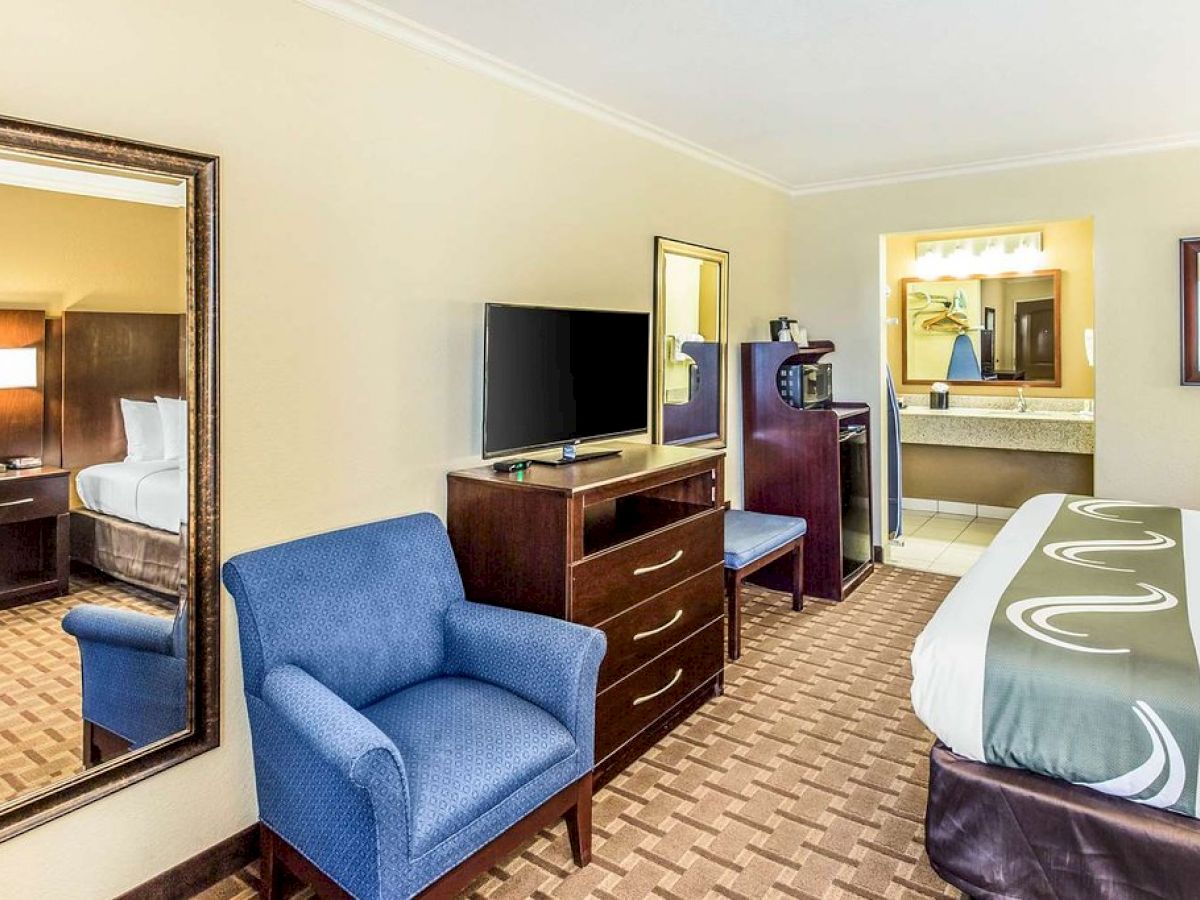 A hotel room with a bed, blue chair, TV on a dresser, large wall mirror, mini fridge, microwave, and sink area in the background.