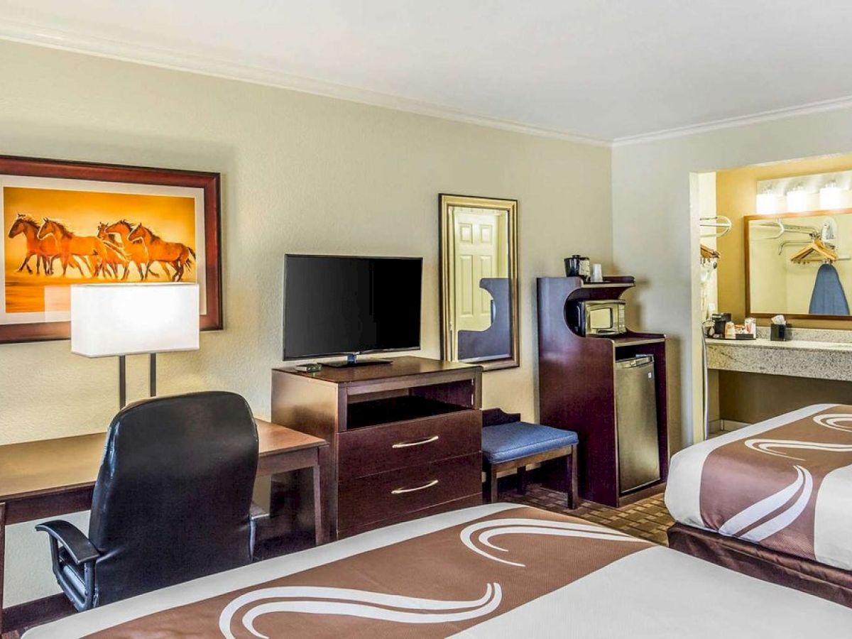 A hotel room features two beds, a desk and chair, a TV, a fridge, microwave, and a vanity area with a mirror and sink.