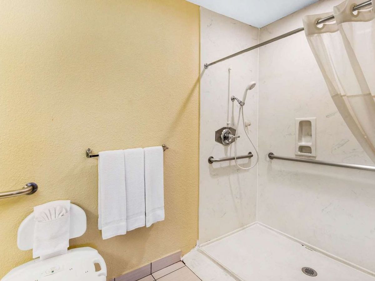 The image shows a bathroom with a walk-in shower, grab bars, a white towel on a rack, and a shower chair.