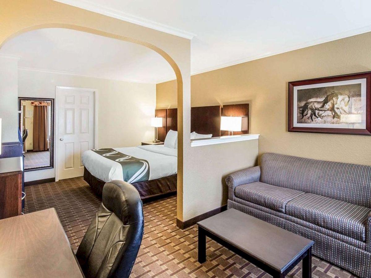 A hotel room with a bed, sofa, desk, office chair, wall art, and a small archway leading to the bed area. The decor is neutral and cozy.